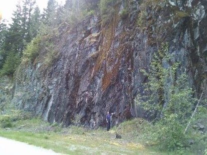 A steep roadside rock face on the claims. Dark exposed rock runs the width of the cut with rust-colored oxidation staining down the center, conifers stand along the top edge, and a person at the base gives scale.